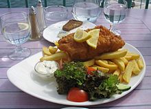 A plate of fish and chips, with salad, dip, lemon slices and a glass of water.