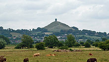 In the distance a small hill with a stone tower on the top. In the foreground flat land with vegetation.
