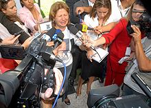 A distraught woman talks into a microphone with her eyes closed, surrounded by reporters. On the front of the woman's white t-shirt there is a printed color portrait of a smiling man. Next to her stands a younger woman with her head down.
