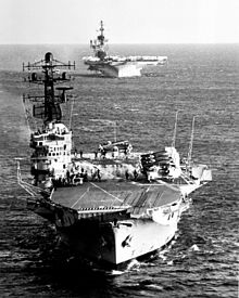 Bow view of an aircraft carrier sailing towards the photogapher. Aircraft with folded wings are sitting on the deck. A second aircraft carrier is following the first, but the upper part of this ship is outside the frame of the photograph.