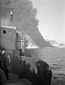 ship at sea with a column of black smoke rising from the mountainous land in the background