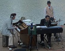 At left man in his thirties plays piano, man in the middle is in his seventies also playing piano, man at right in his twenties or thirties playing a snare drum. On the Mississippi waterfront