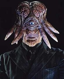 Head to chest portrait of an alien creature with flesh coloured skin. The head is vertically elongated with a small mouth, prominent chin and large, pink exposed brain. A single grey eye is set centrally in the face, with tentacles protruding from the sides of the head. It wears a black shirt and tie with a black pinstripe jacket.