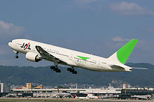 A Boeing 777-200 aircraft in mid air during take-off, with the view of Itami Airport in the background