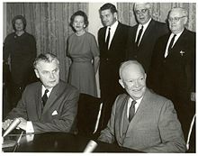 Diefenbaker and a smiling bald man in a suit sit at a table.  Two women and two men stand behind them.
