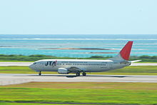 A Boeing 737-400 aircraft painted in Japan Transocean Air livery, travelling along the runway during take-off, with a clear green grass strip in the foreground and a blue sea view in the background