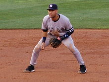 A man in a grey baseball uniform with "New York" written on the front in navy letters and a navy hat with white letters "N" and "Y" interlocking stands ready to field his position.
