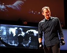 A man wearing a gray shirt, brow pants and black gloves stands in front of a screen, where scenes from Minority Report are being projected.