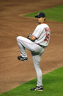 A man in a gray baseball uniform stands on his right foot, ready to deliver a pitch. His right hand is hidden inside of his black baseball glove. The man wears a navy blue baseball cap and has a short goatee.