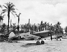 Two propeller aircraft parked on a crushed coral surface. In the background is a coconut plantation