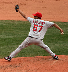 A man in a gray baseball uniform throws a baseball from a dirt mound. He is wearing a red baseball cap. His back is to the camera, and the reverse of the uniform reads "Rodriguez" in red block letters and "57" in a larger red font. His arms and legs are stretched to their limit in four different directions; the baseball is in his right hand, and he is wearing a black baseball glove on his left.
