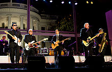 Six male performers playing instruments outdoors, with a background of a white building.