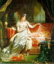 Woman in a white satin dress and tiara sitting on a plush rougey sofa, looking down at a baby lying on the sofa with its eyes closed