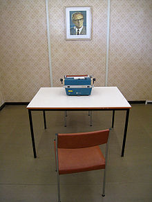 Chair and table with a typewriter on it in a small wallpapered room. A photo portrait of East German leader Erich Honecker is mounted on the rear wall