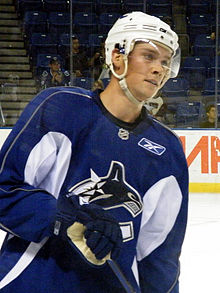 An ice hockey player in his early-twenties standing relaxed on the ice. He wears a blue, visored helmet and a blue jersey with white trim.