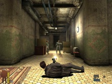 A video game screenshot of a man leaning on the floor while holding a gun in each hand and firing them on another man down a hallway.