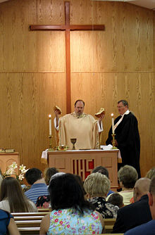 Photo of a service in a church. A United Methodist elder performs the consecration of the bread and wine for Holy Communion. He wears a cream satin robe and stands behind an altar, in front of a large wooden cross. He holds a broken loaf of bread aloft as he prays. On the altar is a large chalice.