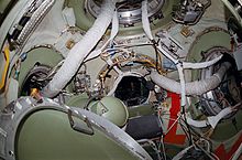 A view of the station's node module, with four open hatches visible. Each hatch is surrounded by a wide green ring, with the node walls coloured white. Numerous ventilation hoses and cables are visible passing between the hatches, and a gyrodyne and hatch cover are seen floating toward the lower left of the image.