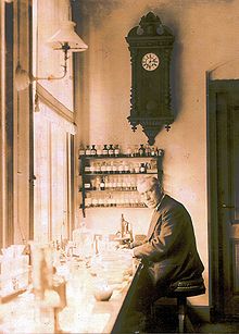 An old, bespectacled man wearing a suit and sitting at a bench by a large window. The bench is covered with small bottles and test tubes. On the wall behind him is a large old-fashioned clock below which are four small enclosed shelves on which sit many neatly labelled bottles.