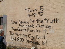 A woman, at the left of the image, is reading a black spray paint message written on a brick wall. The message reads "Team 5 4-19-95 We Search For the truth We Seek Justice. The Courts Require it. The Victims Cry for it. And God Demands it!"