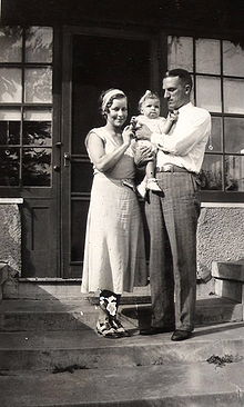 A couple stand on the front steps of a house. The man, aged about 30, dressed in grey flannels and a white shirt, holds a baby girl in his arms and gazes at her. The woman, dressed in a frock and co-respondent shoes from the 1930s stands next to them, touching the baby girl and smiling at the camera. The baby is dressed in a white frilly frock, white shoes and with a white ribbon in her hair.