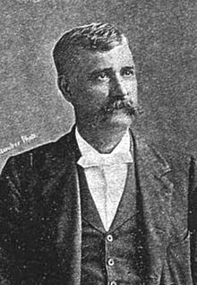 A man in his forties with black hair and a long mustache wearing a white shirt and black coat, facing right