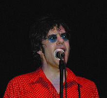 Shoulder high portrait wearing red polka dot shirt and blue sunglasses, sweating, singing into microphone which partly hides his face.