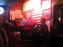 Three musicians, left to right: older man with white hair playing drums, middle-aged man looking into camera, older woman with white hair playing accordion. Walls are covered with an American flag and signs.