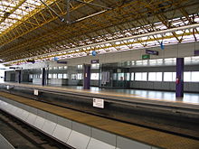 An empty boarding platform well-lit by sunlight streaming from windows on the walls and ceiling from which hangs a yellow latticed net of metal.