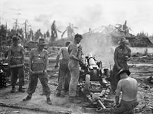 Rear view of an artillery gun mounted on a wheeled carriage. Palm trees, damaged buildings and smoke are visible in front of the gun's barrel. Three shirtless men are working on the gun while another three shirtless men are standing in a line to its left each holding a single artillery shell.