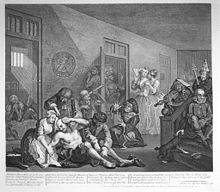 Engraving showing an insane asylum. In the foreground, an almost naked man being shackled and restrained by caretakers. On the stairs to the right, a violinist with sheet music on top of his head, and a man with a dunce cap on. In the background, two well-dressed women have come to view the inmates. Through doors in the background, we can see other mad inmates, all of them almost naked.
