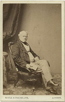 A sepia cabinet photograph of a middle-aged man relaxing in an armchair. He is dressed in mid-nineteenth century clothing, and holding a book in one hand.