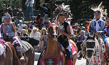 "Colour photograph of Tsuu T'ina children in traditional costume on horseback at a Stampede Parade in front of an audience"