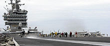 Jet aircraft departing aircraft carrier. A gray-overall aircraft, with blue and yellow fins, has just left the edge of carrier's runway, as evident through the extended landing gear.