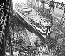 A large warship, still missing most of its superstructure, sits in a dry dock, awaiting its launch. The ship is draped in a large banner and surrounded by crowds of spectators; a huge gantry towers over the ship.
