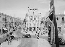  A period photo of street scene. Cars and pedestrians move through an intersection decorated with Union Jacks and Allied flags