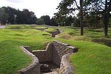 Curved trench lines, preserved in concrete are surrounded by shell craters that are now covered in grass. In the immediate foreground, a small half-destroyer piece of artillery sits in a three walled position that is off of the main trench line.
