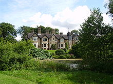 Country house, partially obscured by greenery, viewed from across a pond. The observable frontage comprises five gables, with a turret between the four gables on the left and the rightmost gable.
