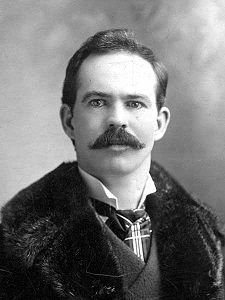Upper body of a man with short hair and a thick moustache.  He is wearing what appears to be a fur coat over a suit.