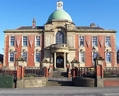 The front of a grand red-brick two-storey structure that is viewed from the ground upwards. It has a complex facade with a central entranceway surrounded by many windows at regular intervals. The roof is crowned by a green cupola and the clear sky appears as a vivid azure.