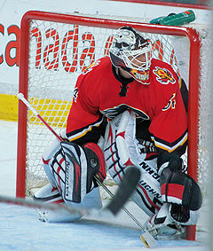 A goaltender in a red uniform and standing in front of his net observes the actions of other players (not seen).