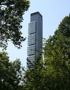A tall, thin building with some slight squarish projections at its higher levels seen from between some trees.