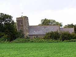 Low building with tiled roof and non vertical square tower, surrounded by trees and with grass in the foreground.