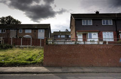 A roadside view of several 20th-century British houses. The houses are set high above the roadside. A grass slope is visible to the lower left of the image, and a tall brick wall to the lower right. A gap in the centre of the image indicates the absence of a single house