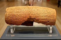 Rear view of a barrel-shaped clay cylinder resting on a stand. The cylinder is covered with lines of cuneiform text