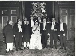 A number of older men in formal dress surround a young woman with a white gown.