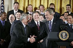 Two men in black suits shake hands smiling, the man on the right standing in front of a podium with the seal of the President of the United States on it.