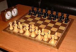 This photo shows a chessboard with pieces set up on both sides, ready to play.  A chess clock is at the side.