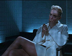 A blonde woman wearing a white jacket, top, and short skirt, her face half in shadow, sitting in an arm chair with her legs crossed. She holds a cigarette to her mouth with her right hand, and raises a lighter with her left. Behind her is dark furniture and the corner of the room, walled with white brick. From between the furniture and walls, unseen, floor-level lights cast a bluish glow over the scene.