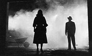 A high-contrast black-and-white image. White fog or smoke fills the background. In the left foreground is the silhouette of a woman wearing a calf-length skirt. To the right, at a further distance, is the silhouette of a man wearing a fedora.